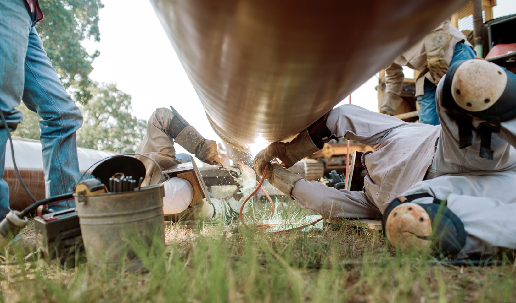 A photograph of two men laying on the ground welding up onto the bottom of a large pipe.