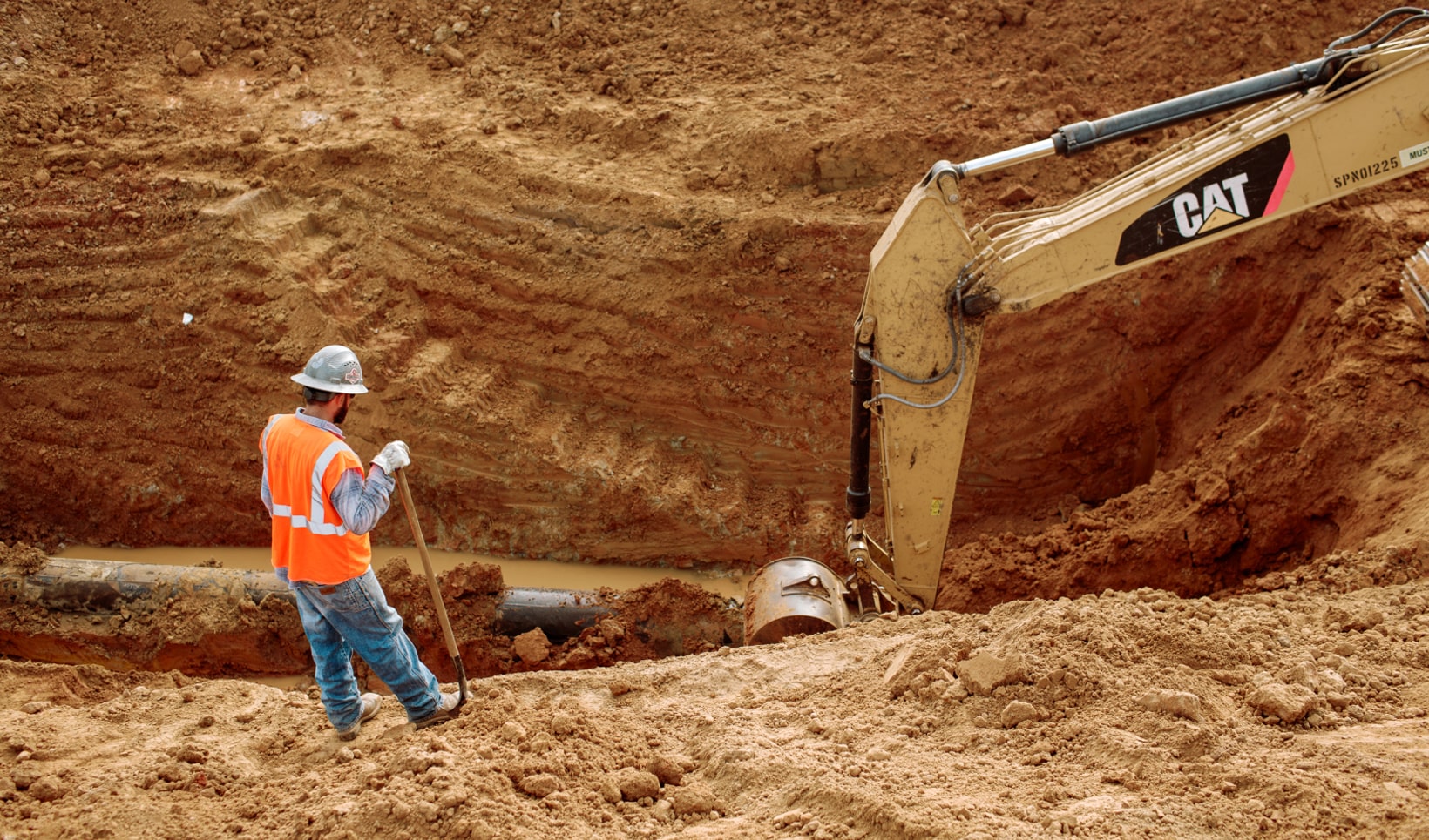 A photograph of a man looking into a large hole being dug out to lay pipe.