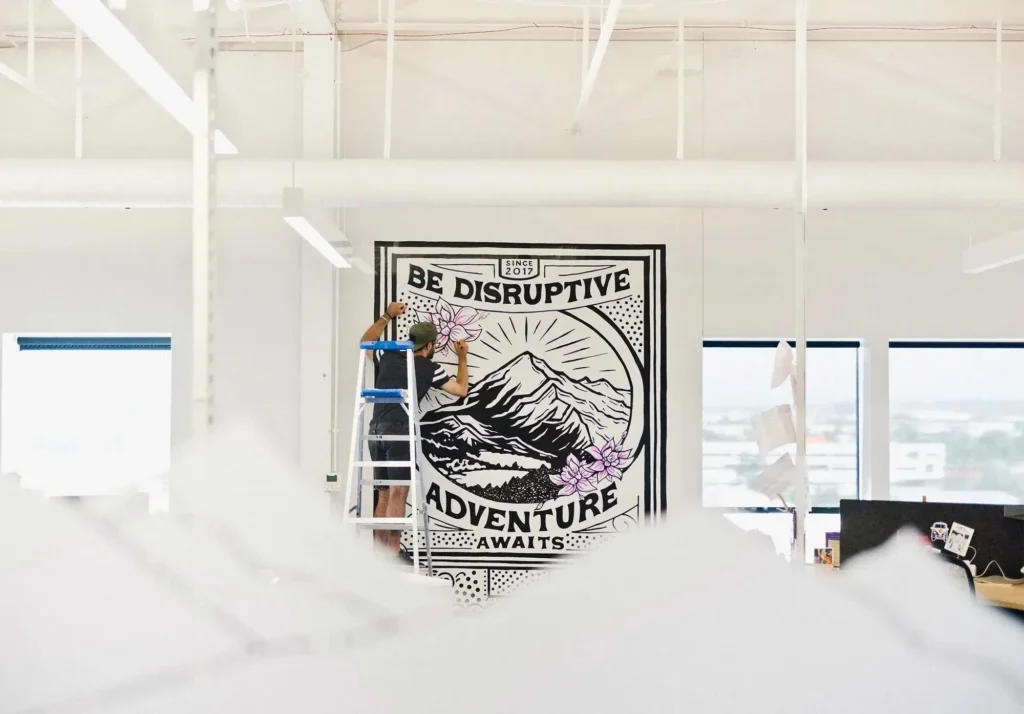 Muralist on a ladder painting a large black ink mountain mural inside an open corporate office.